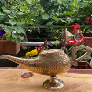 May include: A gold genie lamp with intricate engraved designs and a removable lid. The lamp has a spout and a handle, and it sits on a round base.