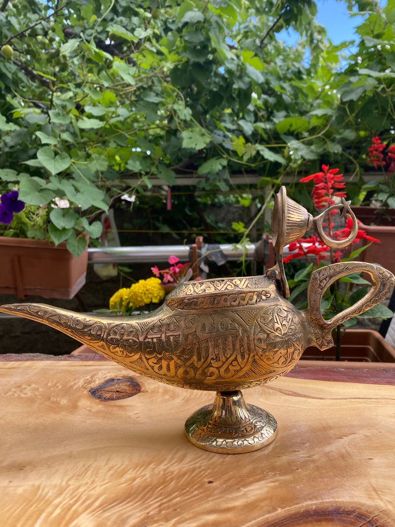 May include: A gold genie lamp with intricate engraved designs and a removable lid. The lamp has a spout and a handle, and it sits on a round base.