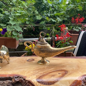 May include: A gold genie lamp with intricate designs sits on a wooden table. The lamp has a curved spout and a handle in the shape of a crescent moon.