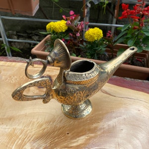 May include: A gold genie lamp with intricate details and a hinged lid. The lamp is sitting on a wooden surface.