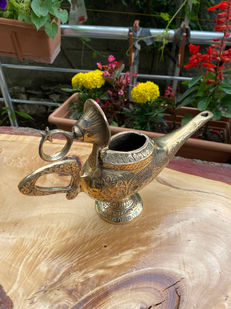 May include: A gold genie lamp with intricate details and a hinged lid. The lamp is sitting on a wooden surface.