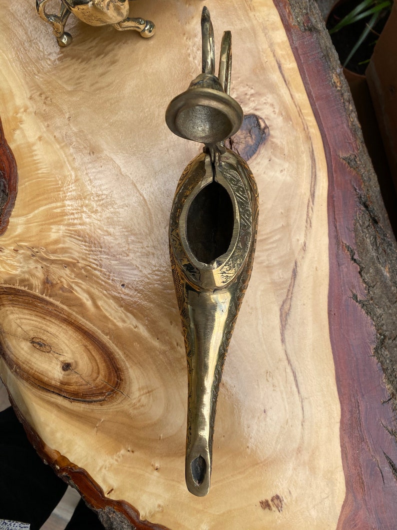 May include: A gold metal genie lamp with intricate details and an open lid. The lamp is on a wooden surface.