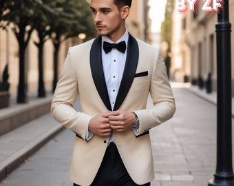 May include: A man wearing a beige tuxedo jacket with a black satin lapel and a black bow tie. The jacket has a single button closure and a pocket on the left side. The man is standing in front of a building with a brick facade.