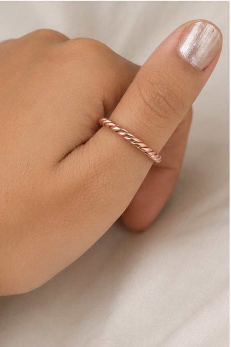 May include: A thin rose gold ring with a twisted rope texture. The band features a continuous spiral design that wraps around the entire circumference of the ring, creating a uniform and detailed metallic surface with a polished finish.