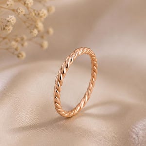 May include: A thin, rose gold ring featuring a continuous twisted rope design. The band has a polished, reflective surface that catches the light, creating a uniform, spiraling texture around the entire circumference of the circular metal ring.