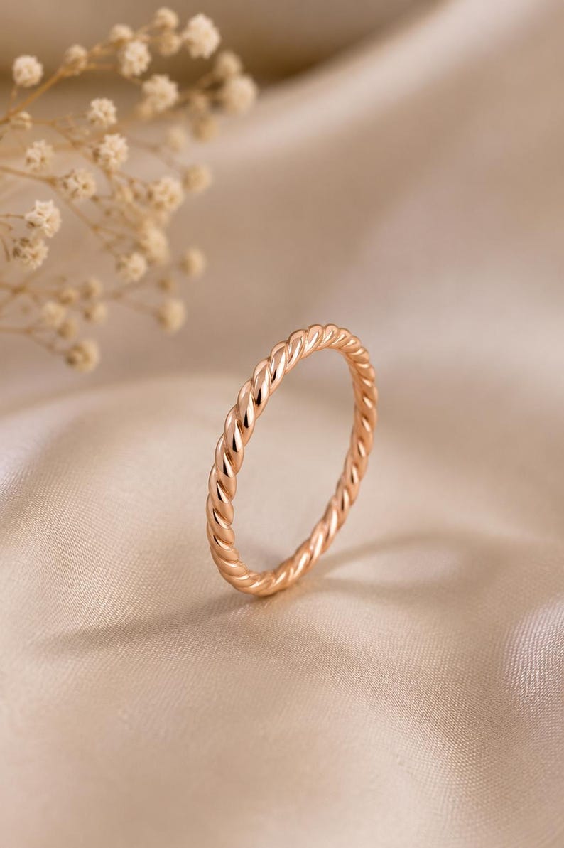 May include: A thin, rose gold ring featuring a continuous twisted rope design. The band has a polished, reflective surface that catches the light, creating a uniform, spiraling texture around the entire circumference of the circular metal ring.