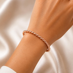 Op de afbeelding: Een roségoudkleurige armband met een doorlopend gedraaid touwontwerp. Het metaal heeft een gepolijst, reflecterend oppervlak dat het licht opvangt, waardoor een uniforme metalen uitstraling rondom de gehele omtrek van de cirkelvormige band ontstaat.