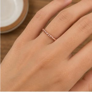 May include: A thin rose gold ring featuring a twisted rope design. The band has a polished metallic finish and a continuous spiral pattern that wraps around the entire circumference of the ring, creating a textured look on the finger.