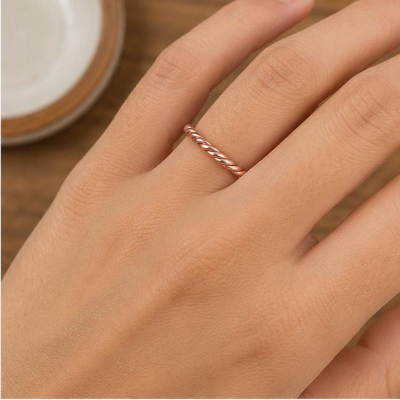 May include: A thin rose gold ring featuring a twisted rope design. The band has a polished metallic finish and a continuous spiral pattern that wraps around the entire circumference of the ring, creating a textured look on the finger.