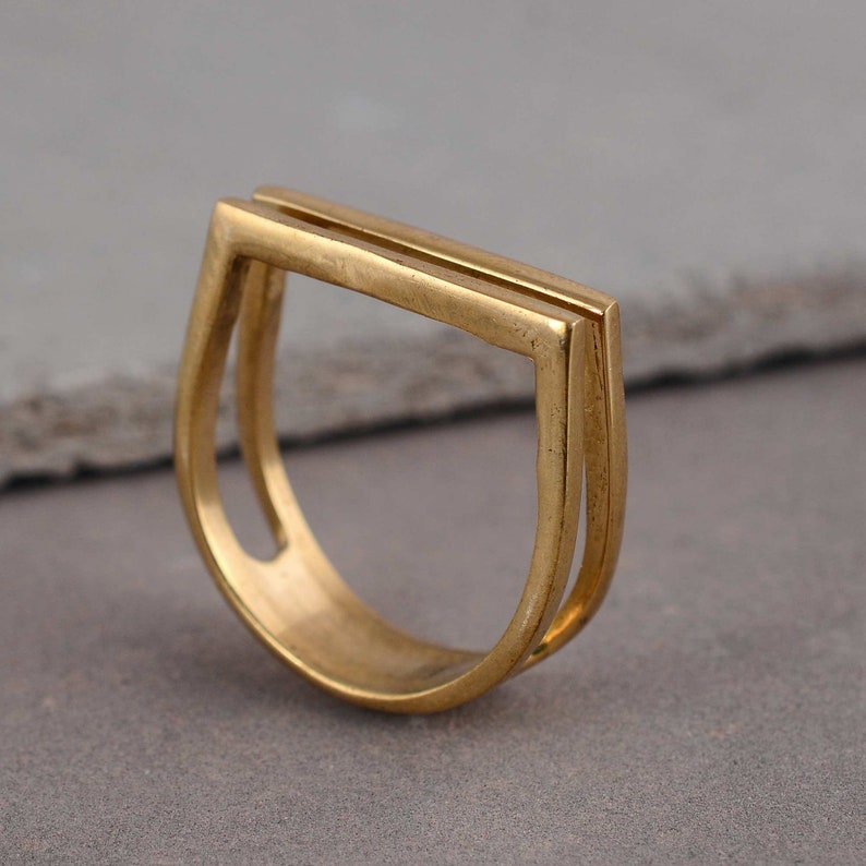 May include: A gold-toned ring with a double-layered, rectangular design. The ring is open on one side and has a rounded shape.