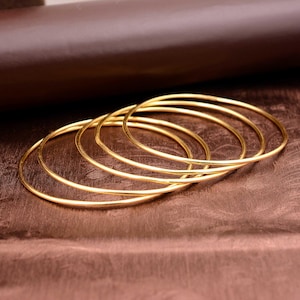 May include: Several gold-coloured bangles, overlapping slightly. The bangles are thin and circular, reflecting light. The background is a gradient of brown and bronze tones, providing a warm contrast to the jewellery.