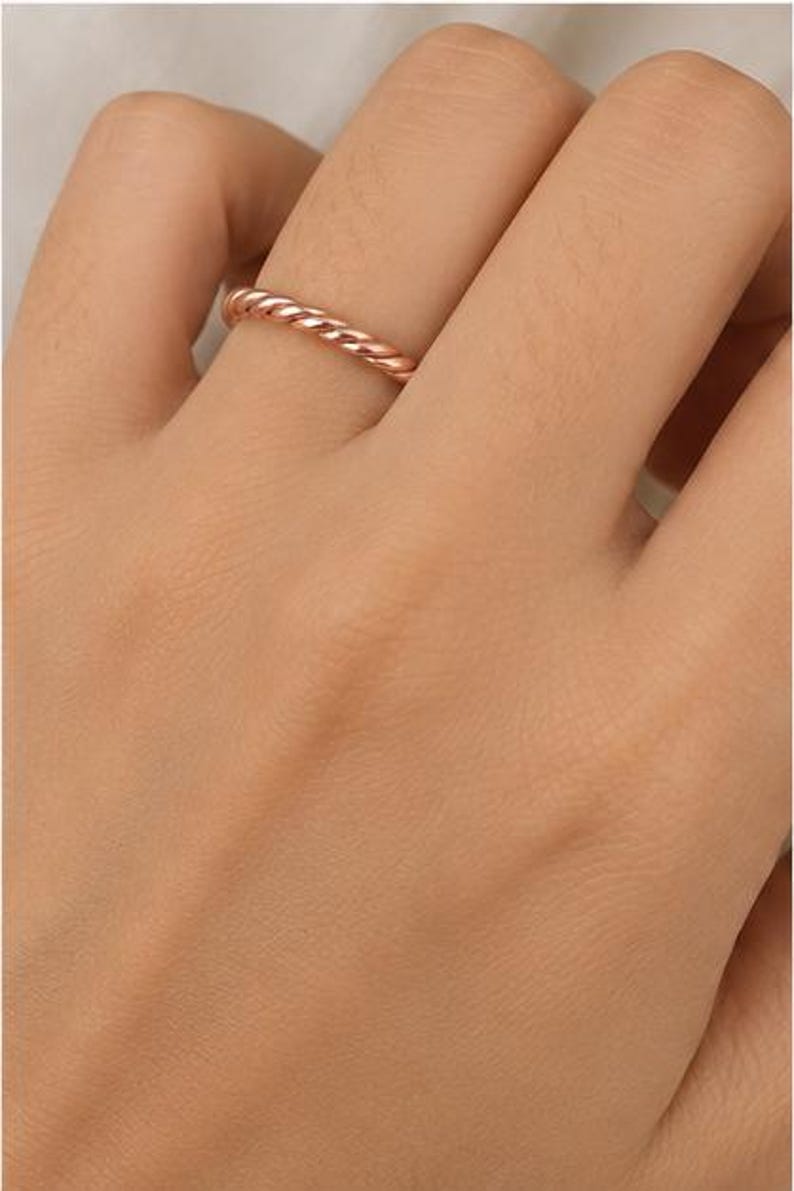 May include: A rose gold ring featuring a twisted rope design. The band has a polished, metallic finish with a continuous spiral pattern that wraps around the entire circumference of the ring, creating a textured appearance on the surface.
