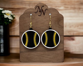 Könnte beinhalten: Ein Paar Ohrringe mit schwarzen und weißen Softball-Designs mit gelben Nähten. Die Ohrringe werden auf einem Holzständer präsentiert.