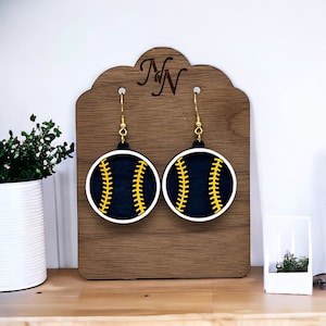 May include: A pair of wooden earrings shaped like baseballs with black and yellow stitching. The earrings are displayed on a wooden stand with the letters "MN" engraved on it.