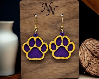 May include: Purple and gold glitter paw print earrings on a wooden display stand. The earrings are shaped like a paw print with a gold outline and purple glitter inside.