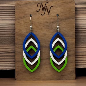 May include: A pair of layered, teardrop-shaped earrings in blue, white, and green, hanging from silver hooks. The earrings are displayed on a wooden plaque with the initials "NN" engraved at the top.