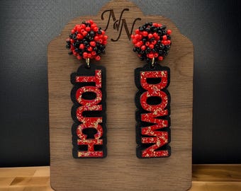 Könnte beinhalten: Ein Paar Statement-Ohrringe. Jeder Ohrring hat oben eine Ansammlung roter und schwarzer Perlen, mit einem schwarzen Ausschnitt, der "TOUCH" und "DOWN" in Rot mit goldenen Glitzerakzenten schreibt. Die Ohrringe werden auf einem Holzständer präsentiert.