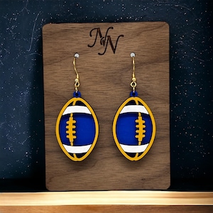 May include: A pair of football-shaped earrings. Each earring features a blue center with white and yellow accents, and a gold-colored hook. The earrings are displayed on a wooden plaque with the initials "NN" engraved on it.