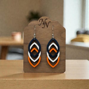 May include: A pair of layered, teardrop-shaped earrings in navy blue, white, and orange. The earrings are displayed on a wooden stand with the initials "NN" carved at the top. The earrings have a modern, geometric design.