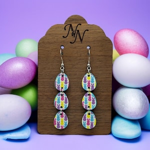 May include: A pair of dangle earrings with three teardrop-shaped wooden charms. Each charm is decorated with a pattern of colorful Peeps marshmallow bunnies. The earrings are displayed on a wooden stand with the letter "N" carved into it.