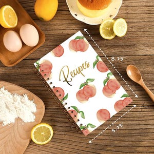 May include: A spiral-bound recipe notebook with a peach pattern and the word "Recipes" in gold script. The notebook measures 8.75" x 5.75". Ingredients like eggs, lemons, and flour are arranged around the notebook on a wooden surface.