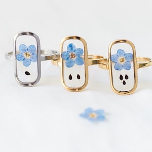 May include: Three rings with rectangular settings, each featuring a small blue flower. One ring has a silver band, while the other two have gold bands. Scattered around the rings are small blue flowers, creating a delicate and floral theme.