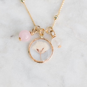 May include: A gold chain necklace with a delicate pendant featuring a pressed flower encased in resin. The necklace also includes small pink and white beads.