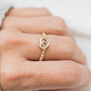 May include: A gold ring featuring a small heart-shaped charm with a clear centre and a delicate beaded band. The ring is worn on a finger, showcasing the metallic gold finish and the simple, open-frame heart design that sits centred on the band.
