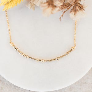 May include: A gold-toned chain necklace featuring a central segment of small, alternating round beads and thin cylindrical bars arranged in a sequence, creating a delicate and refined piece of jewelry with a polished metallic finish.