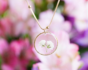May include: A gold chain necklace with a round pendant featuring two small white flowers pressed inside clear resin.