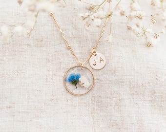 May include: A gold-toned necklace featuring a circular pendant containing a pressed blue and white flower. Attached to the chain is a small, round gold-toned charm engraved with the letters JA. The chain has a delicate link design with small beads.