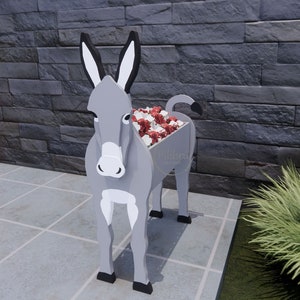 May include: A gray donkey-shaped planter filled with red and white flowers. The planter has black ears, hooves, and tail. The donkey is standing on a tiled surface in front of a dark gray brick wall. The planter has the text "Pilibra Designs".