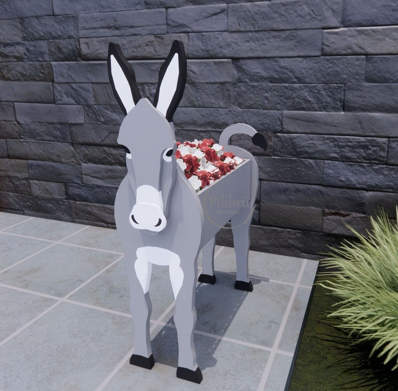 May include: A gray donkey-shaped planter filled with red and white flowers. The planter has black ears, hooves, and tail. The donkey is standing on a tiled surface in front of a dark gray brick wall. The planter has the text "Pilibra Designs".