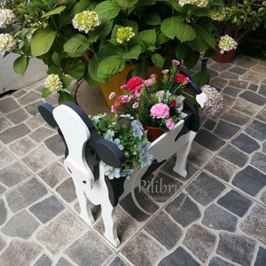 May include: A black and white cow-shaped planter with pink accents. The planter is filled with pink, white, and blue flowers. The planter is sitting on a stone patio.