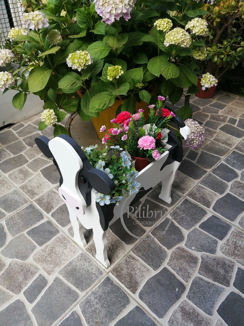 May include: A black and white cow-shaped planter with pink accents. The planter is filled with pink, white, and blue flowers. The planter is sitting on a stone patio.