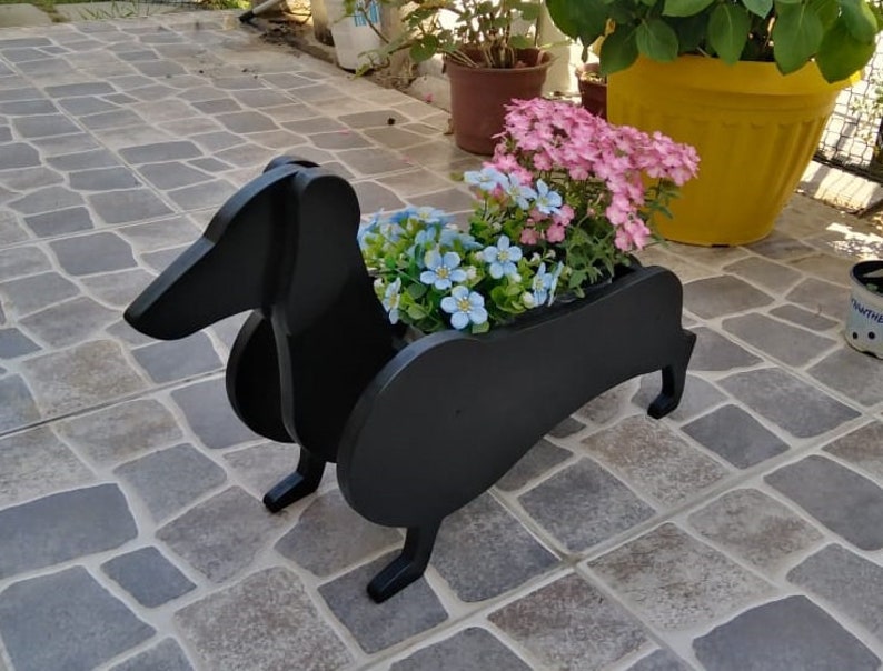 May include: Black wooden dachshund planter with pink and blue flowers. The planter is sitting on a stone patio.