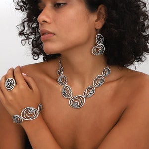 Può includere: Un set di collana, braccialetto e orecchini in argento con un design a spirale circolare. I gioielli sono in metallo e hanno una finitura lucida.