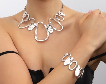 May include: A silver-toned necklace and bracelet set. The necklace features an abstract design with irregular shapes and a chain closure. The matching bracelet has a similar design. The jewelry is worn against a black top.