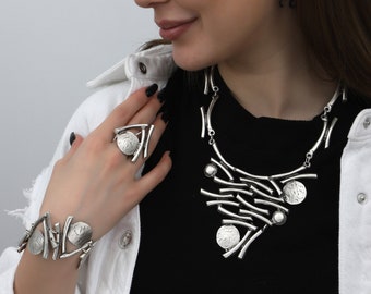 May include: A silver jewelry set featuring a necklace, bracelet, and ring. The necklace and bracelet have a linear design with circular accents. The ring has a similar design. The jewelry is worn with a black top and white jacket.