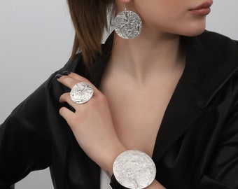 May include: A silver jewelry set featuring a textured, circular design. The set includes a ring, earrings, and a bracelet. The bracelet has a black band. The earrings are dangling. The jewelry has a hammered metal appearance.