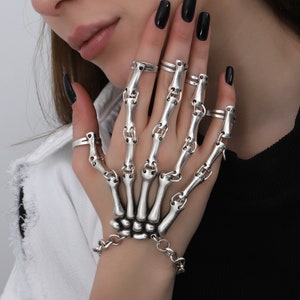 May include: A silver skeleton hand ring with multiple connected rings. The ring is worn on the index, middle, and ring fingers of a hand.