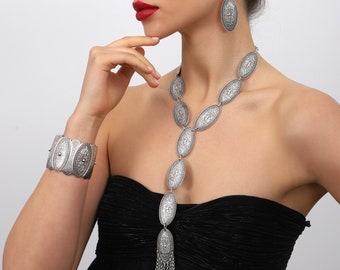 May include: A silver-toned jewelry set featuring an oval pendant necklace with chain tassels, matching earrings, and a cuff bracelet. The jewelry has an ornate, engraved design and is worn with a black strapless dress.