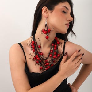 May include: A woman wearing a black dress with a red berry necklace and matching earrings. The necklace is made of black string with red berries attached to it. The earrings are also made of black string with red berries attached to them.