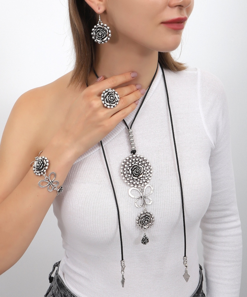 May include: A silver-toned jewelry set featuring a long necklace with a large rose pendant, a smaller rose pendant, and a butterfly charm. The set also includes a matching ring and bracelet with rose and butterfly designs.