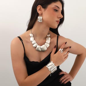 Peut inclure: Un ensemble de bijoux argentés comprenant un collier, des boucles d'oreilles, un bracelet et une bague. Le collier et les boucles d'oreilles ont de grands carrés irréguliers. Le bracelet a un design superposé et la bague une forme de demi-lune. Les bijoux sont portés avec une robe noire.