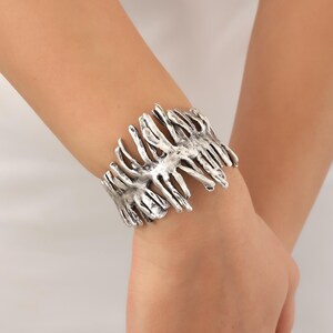 May include: Silver-toned cuff bracelet with a textured, organic design resembling branches or a jagged edge. The bracelet is open and adjustable.