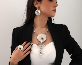 May include: A silver necklace with three large, round, hammered metal pendants. The necklace is worn with a silver ring with a similar design and silver earrings with a single round pendant.