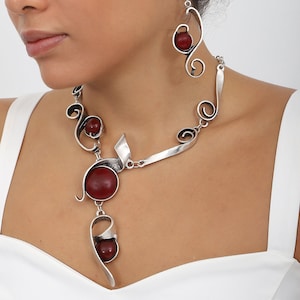 Peut inclure: Un collier et des boucles d'oreilles en argent avec des accents de pierres précieuses rouges. Le collier présente un design long et asymétrique avec un motif en spirale et trois pierres précieuses rondes. Les boucles d'oreilles sont de style pendantes avec une seule pierre précieuse ronde.