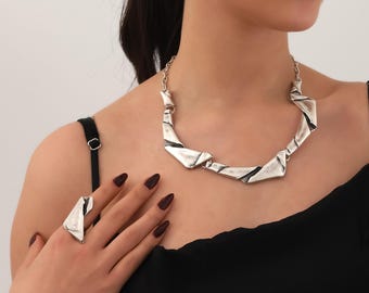 May include: A silver necklace and ring set with geometric shapes. The necklace is made of a chain with a series of connected, angular, silver pieces. The ring is a single, angular, silver piece.