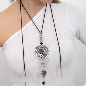 May include: A silver necklace with a large, round pendant featuring a floral design. The pendant is attached to a black cord and has a smaller flower charm hanging below it.
